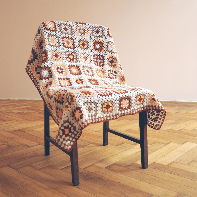 A wooden chair draped with a colorful granny square blanket in warm tones, set against a light-colored wall and wooden floor.