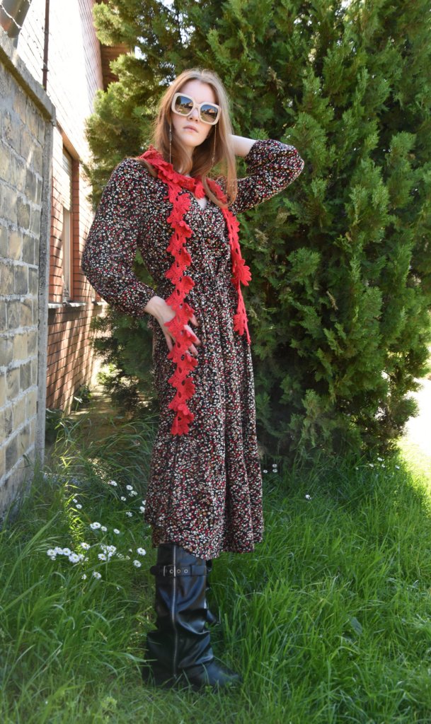 A woman stands outdoors next to greenery, wearing a floral dress paired with a red scarf and large sunglasses. She is striking a pose with one hand in her hair and the other at her waist.